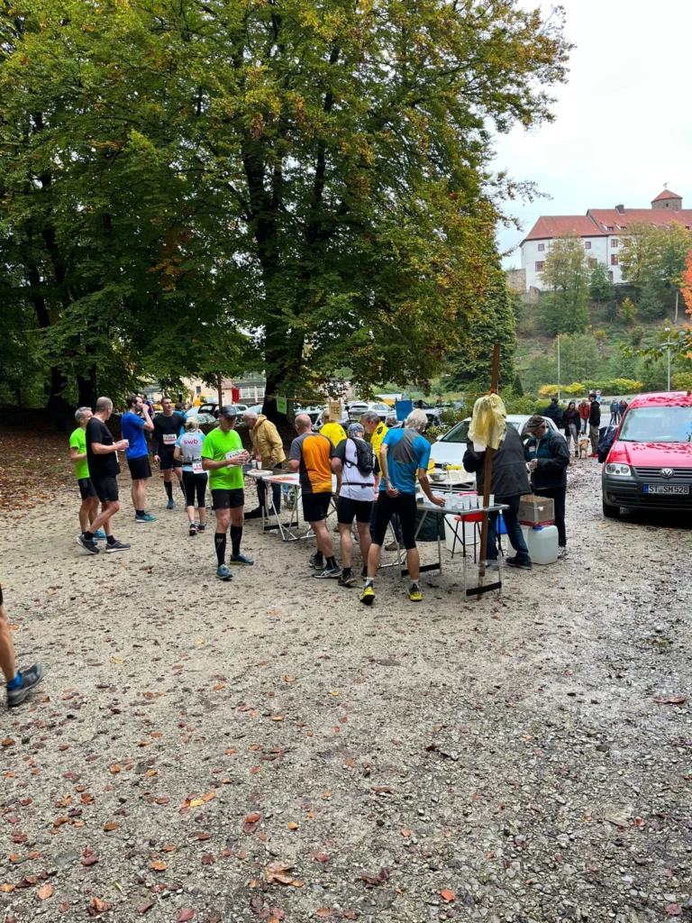 Teilnehmer eines Laufwettbewerbs an einer Verpflegungsstation im Park, mit Bäumen und einem Gebäude im Hintergrund.