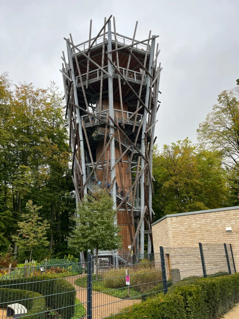 Moderner Holzturm in einem Waldgebiet, umgeben von Bäumen und eingezäuntem Gartenpfad bei bewölktem Himmel.