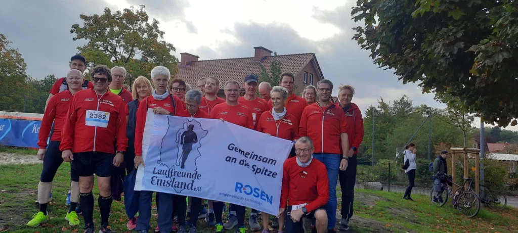 Laufgruppe posiert mit Banner vor herbstlicher Kulisse, trägt rote Jacken mit Vereinslogo Lauffreunde Emsland e.V..