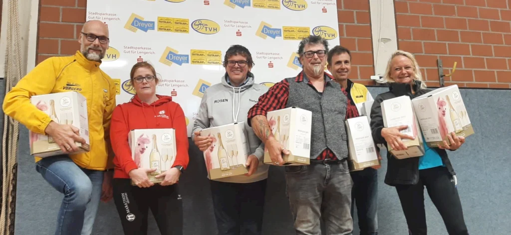 Sechs Personen halten Geschenkverpackungen mit Sekt vor einer Sponsorenwand bei einer Veranstaltung.