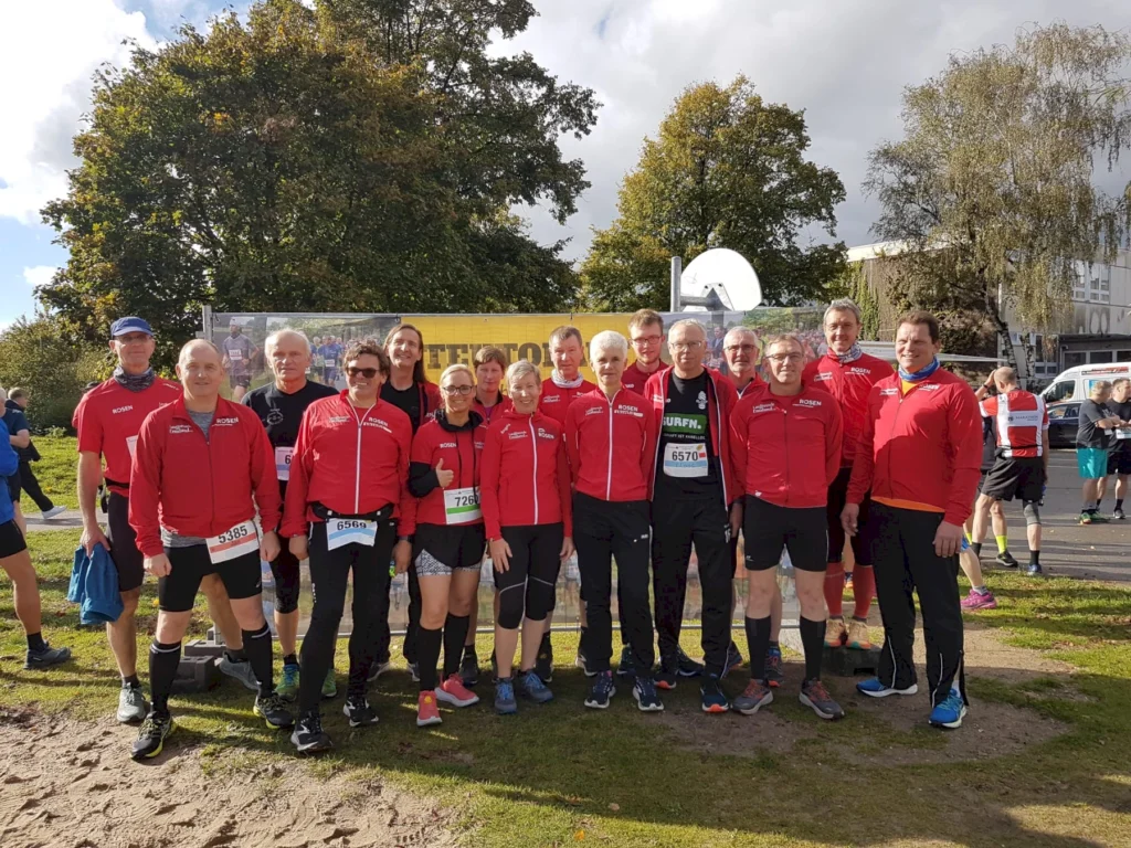 Gruppe von Läufern in roten Trainingsanzügen beim Outdoor-Wettbewerb, sonniger Tag, grüne Bäume im Hintergrund.