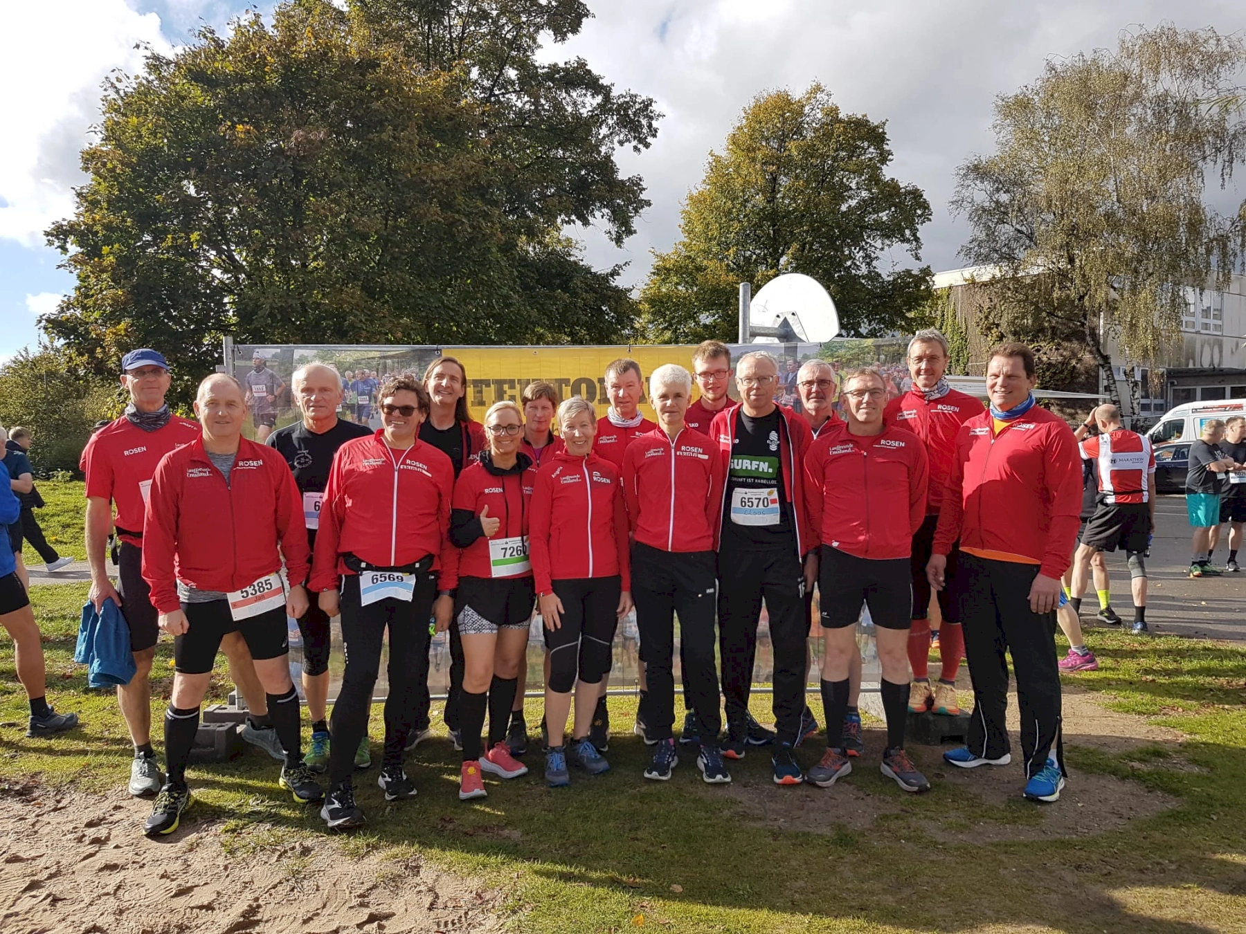 Gruppe von Läufern in roten Trainingsanzügen beim Outdoor-Wettbewerb, sonniger Tag, grüne Bäume im Hintergrund.