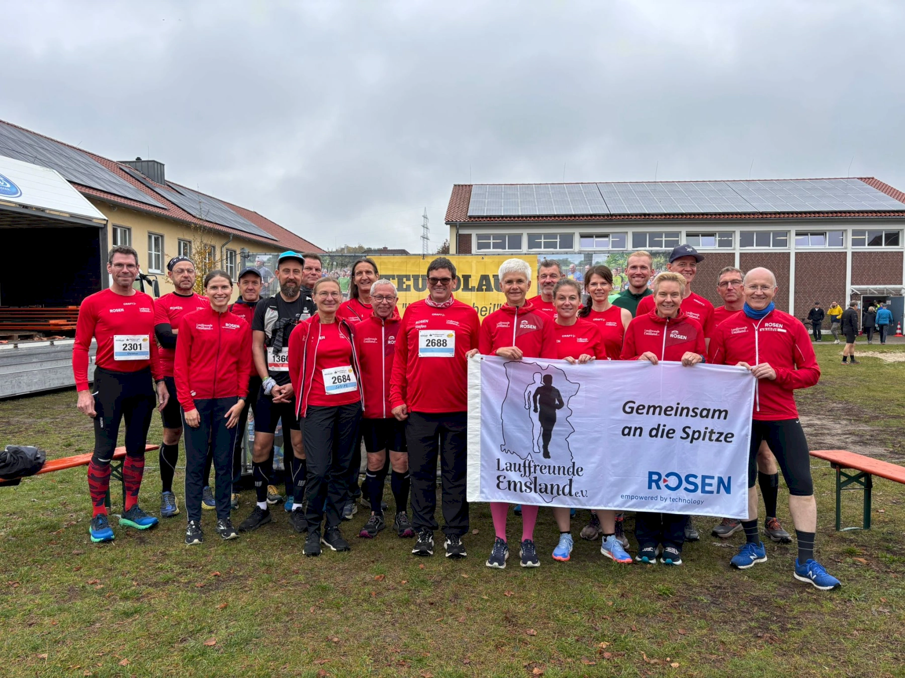 Läufergruppe der Lauffreunde Emsland mit Banner beim Wettkampf auf einer Wiese vor einem Gebäude, gemeinsam in Rot gekleidet.