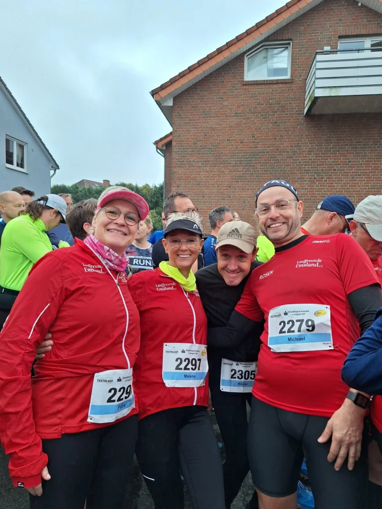 Gruppe von Läufern in roten Jacken mit Startnummern, lächelnd vor einem Haus beim Lauf-Event.