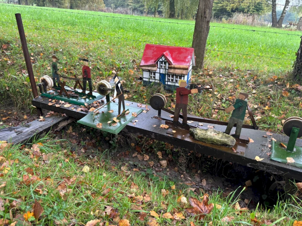 Holzfiguren auf Wasserrad mit Miniaturhaus im Hintergrund, umgeben von herbstlichem Laub und grünem Feld.