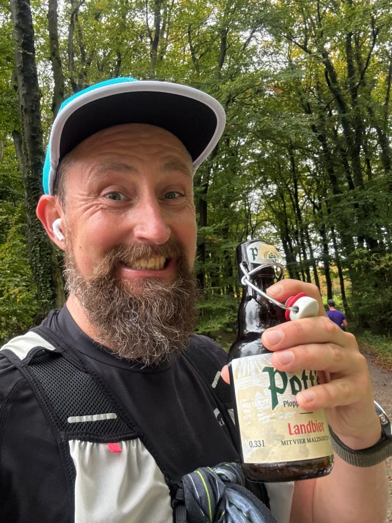 Mann im Wald mit blaubeiger Kappe hält eine Flasche Bier in der Hand und lächelt in die Kamera.