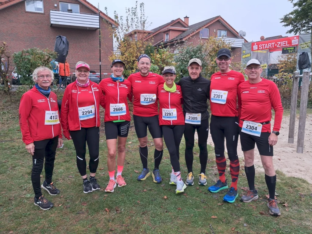Läufergruppe in roten Trikots steht lächelnd beim Start-Ziel-Bereich eines Herbstlaufes vor Backsteinhäusern.