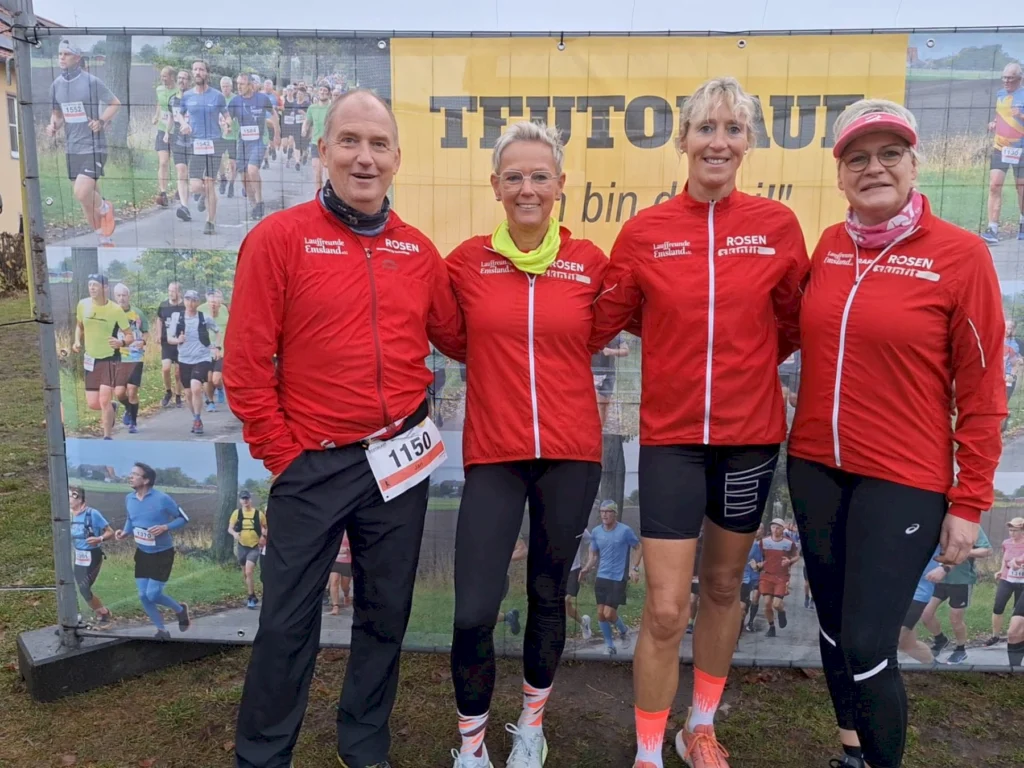 Vier Teilnehmende in roten Jacken posieren vor einem Laufplakat bei einem Outdoor-Lauf-Event.