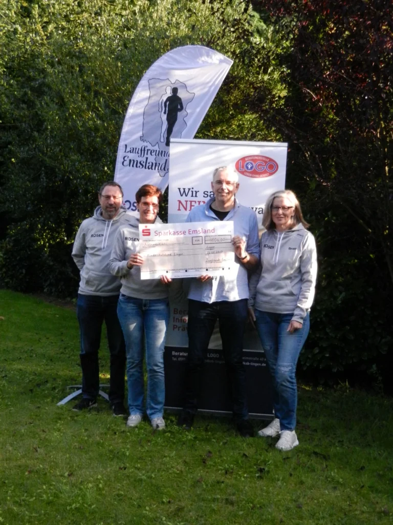 Vier Personen präsentieren einen großen Scheck im Freien, mit Werbebannern im Hintergrund.