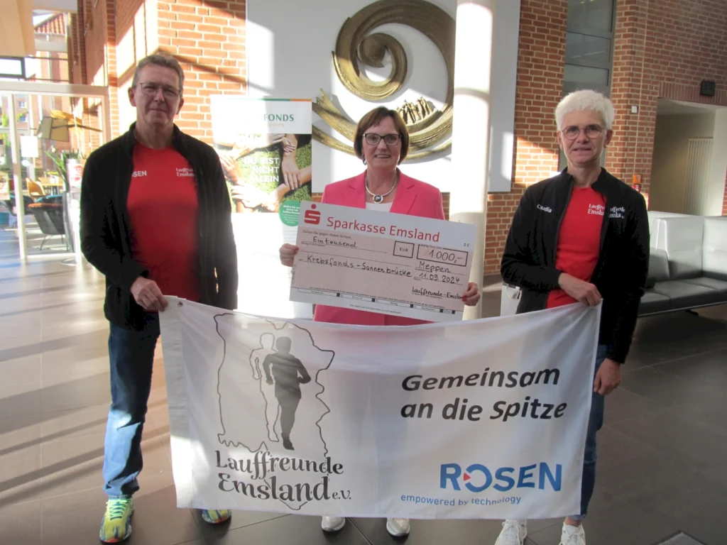 Drei Mitglieder der Laufgruppe Emsland e.V. halten einen Scheck über 1.000 Euro für die Krebsfonds Sonnenbrücke in Meppen. Sie stehen in einem modernen Innenraum und präsentieren das Banner der Laufgruppe.