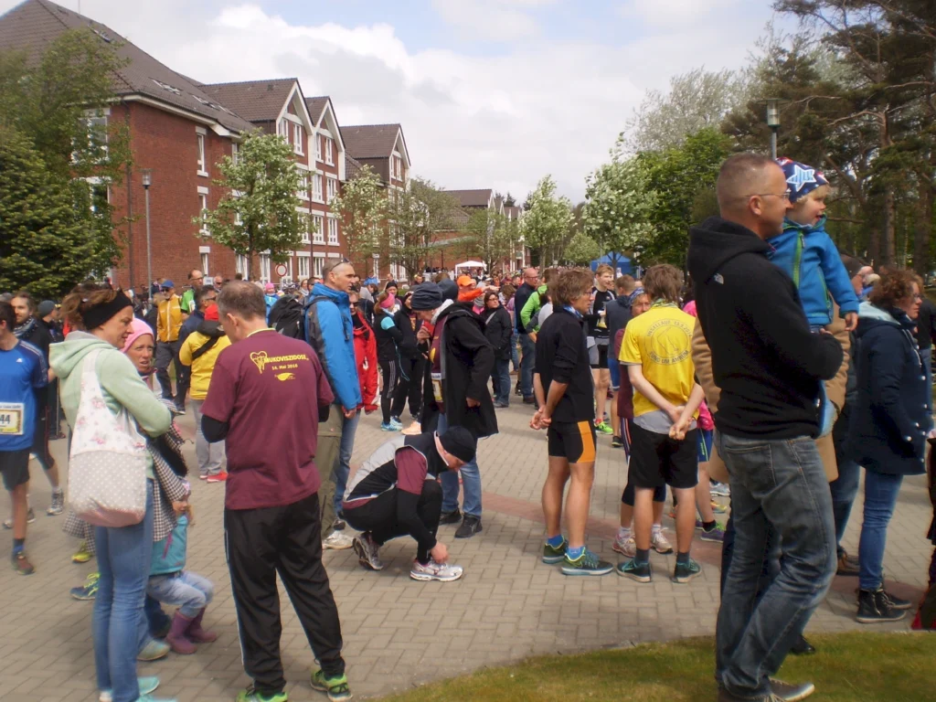 Menschenmenge bei einem Laufereignis, Teilnehmer in Sportkleidung, verschiedene Altersgruppen, fröhliche Stimmung und Vorbereitungen im Freien, umgeben von Bäumen und Wohngebäuden.