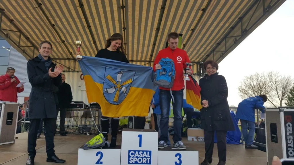 Siegerehrung bei einem Sportevent mit drei Gewinnern auf dem Podium, die Trophäen halten und eine Flagge präsentieren, umgeben von applaudierenden Zuschauern.