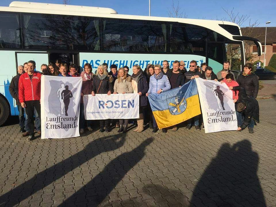 Gruppenfoto von Mitgliedern der Laufgruppe "Lauffreunde Emsland" vor einem Bus, mit Flaggen der Gruppe und des Unternehmens ROSEN, an einem sonnigen Tag.