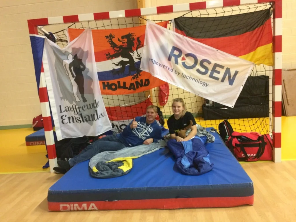 Zwei Personen sitzen auf einer blauen Matratze in einer Sporthalle, umgeben von Fahnen der Lauffreunde Emsland, Holland und Deutschland. Sie entspannen sich in Schlafsäcken und zeigen Daumen hoch. Im Hintergrund sind Sportgeräte und Taschen sichtbar.