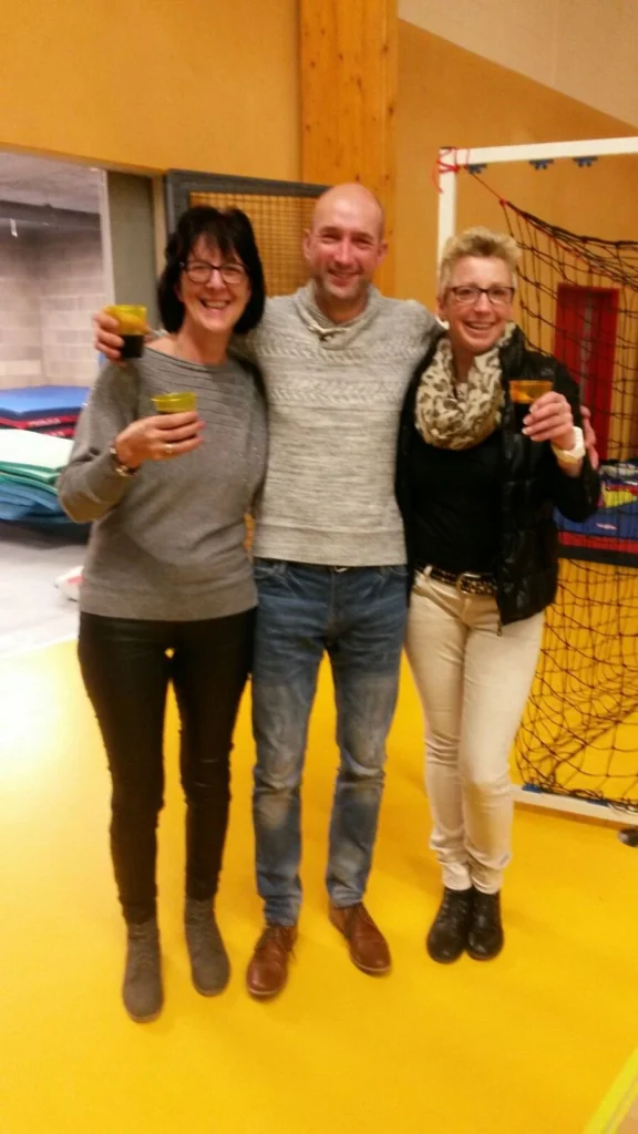 Drei Personen stehen in einer Sporthalle und halten Getränke in den Händen. Die Szene vermittelt eine freundliche, gesellige Atmosphäre. Im Hintergrund sind Sportgeräte sichtbar, die auf eine aktive Veranstaltung hinweisen.