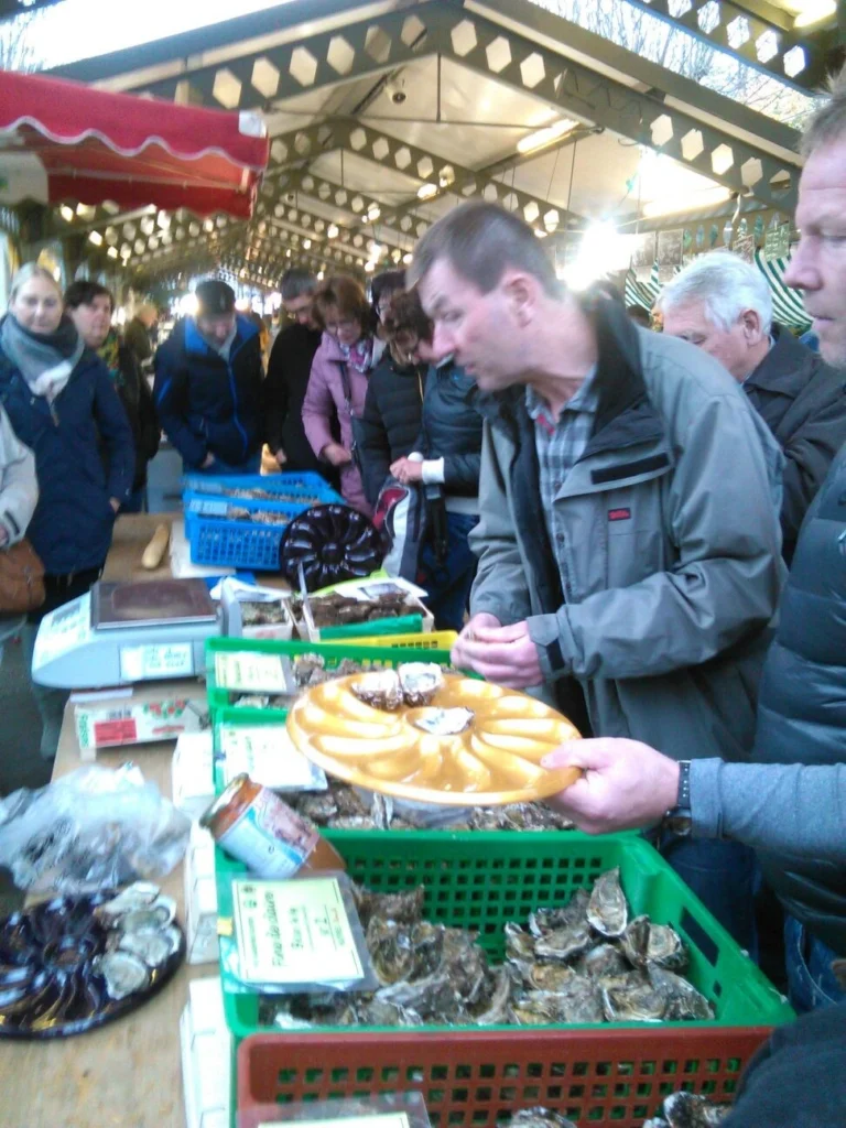 Marktstand mit frischen Austern und Meeresfrüchten, umgeben von interessierten Kunden. Menschen betrachten die Auswahl, während ein Verkäufer eine Servierplatte mit Austern hält. Der Markt ist beleuchtet und bietet eine einladende Atmosphäre für Feinschmecker.
