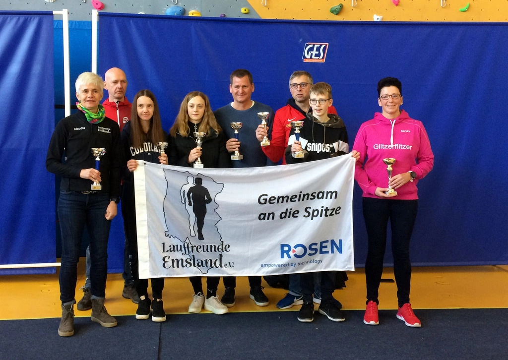 Gruppe von Läufern und Unterstützern der Laufgruppe Emsland e.V. bei einer Siegerehrung, halten Pokale und eine Banner mit dem Slogan „Gemeinsam an die Spitze“.