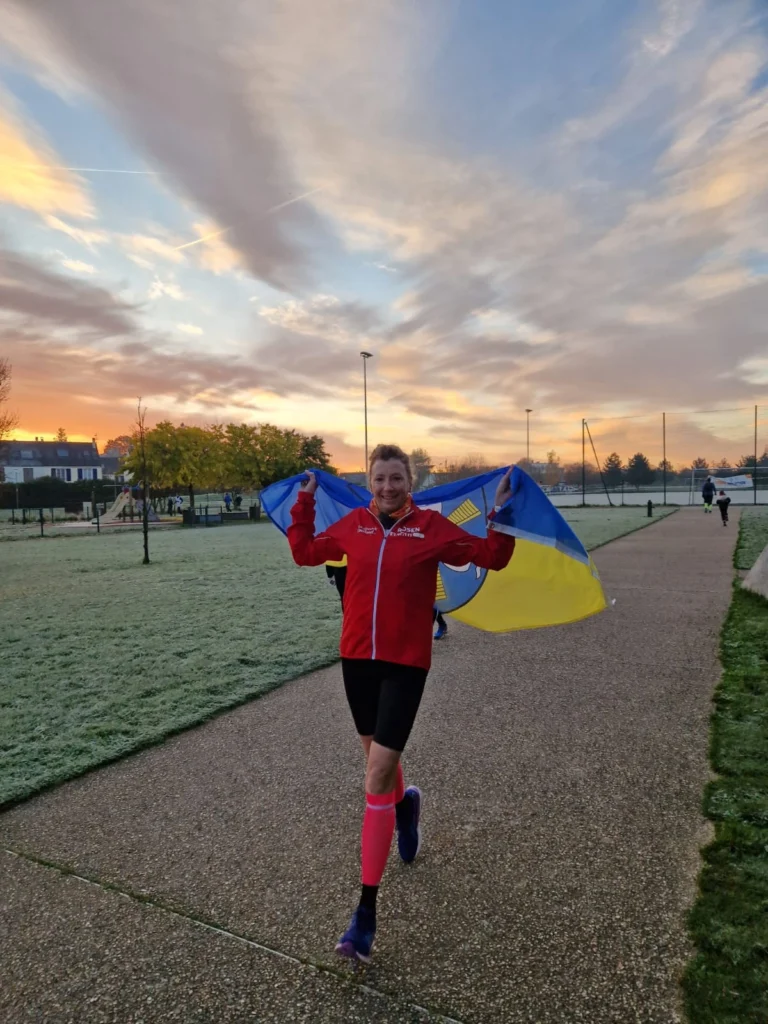 Läuferin mit einer ukrainischen Flagge in der Hand, die auf einem frostigen Weg im Freien joggt, während die Sonne im Hintergrund aufgeht und den Himmel in sanften Farben erleuchtet.