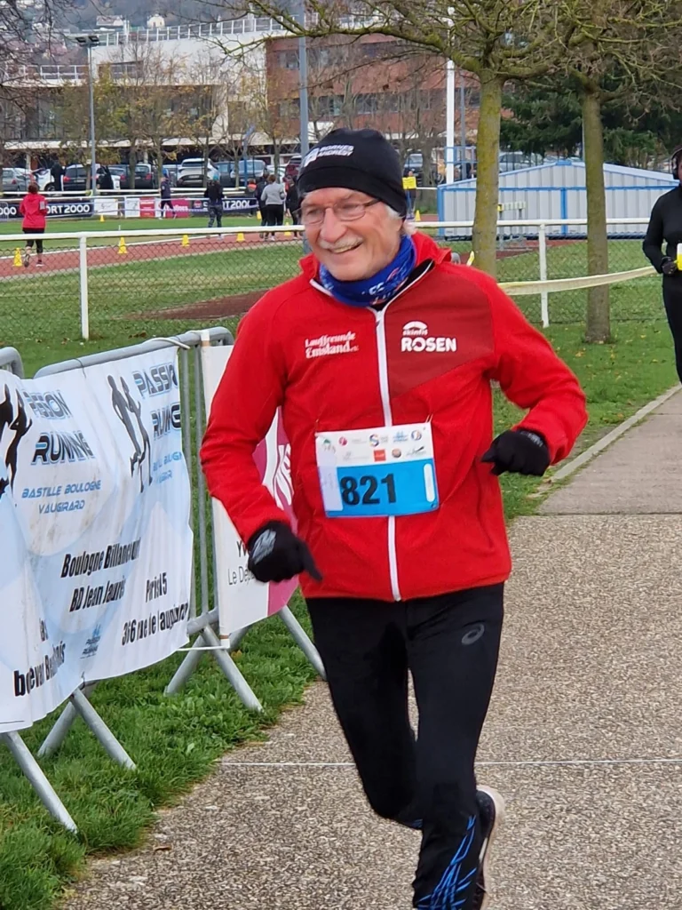 Ein älterer Läufer in einem roten Trainingsanzug mit der Nummer 821 lächelt, während er einen Wettlauf auf einer Laufstrecke absolviert. Im Hintergrund sind andere Teilnehmer und Werbebanner zu sehen.
