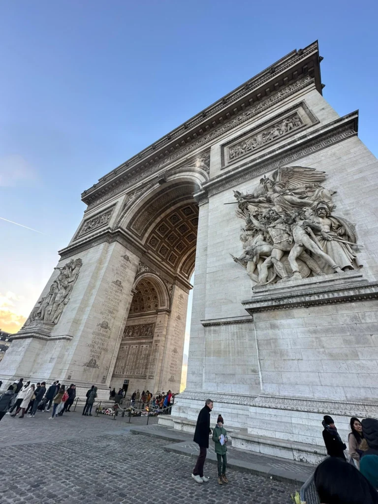 Blick auf den Arc de Triomphe in Paris mit Besuchern, die die monumentale Struktur und die detaillierten Skulpturen bewundern. Der Himmel ist klar und die Atmosphäre lebhaft.