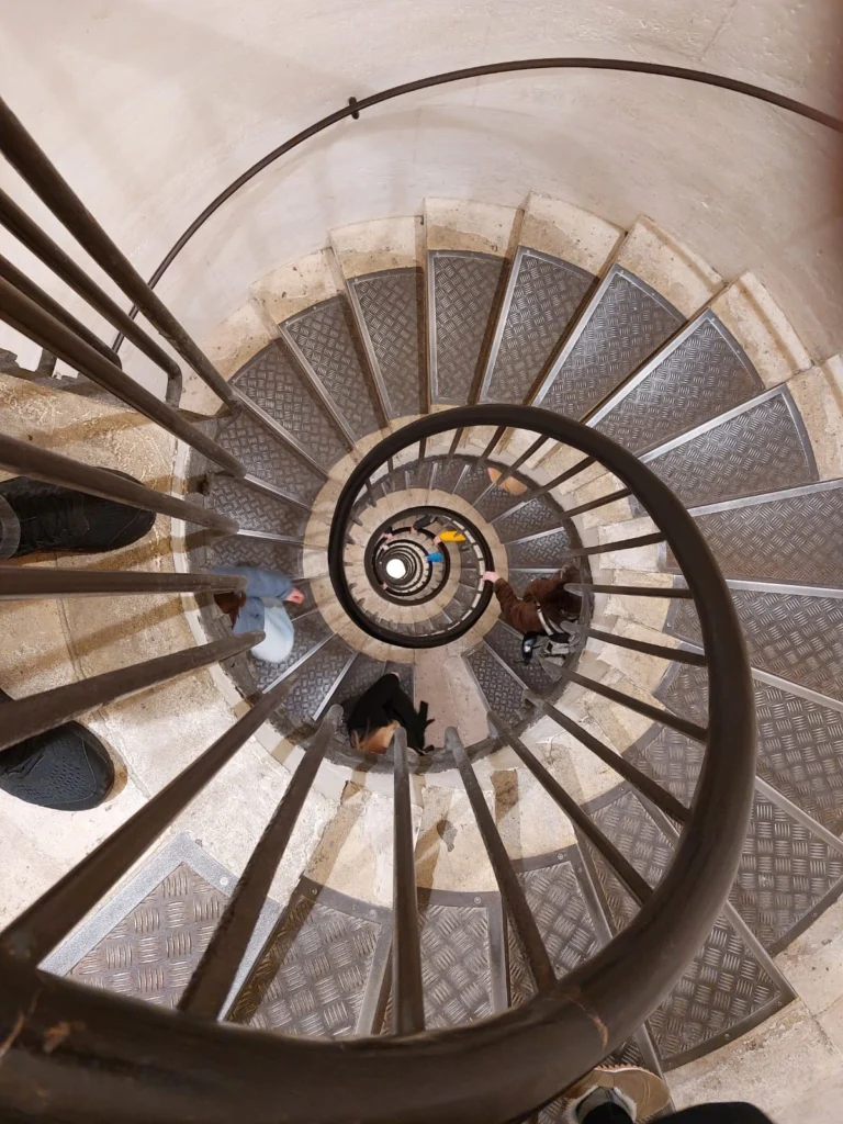 Blick von oben auf eine spiralförmige Treppe mit Metallgeländer und rutschfestem Bodenbelag, die von mehreren Personen genutzt wird.