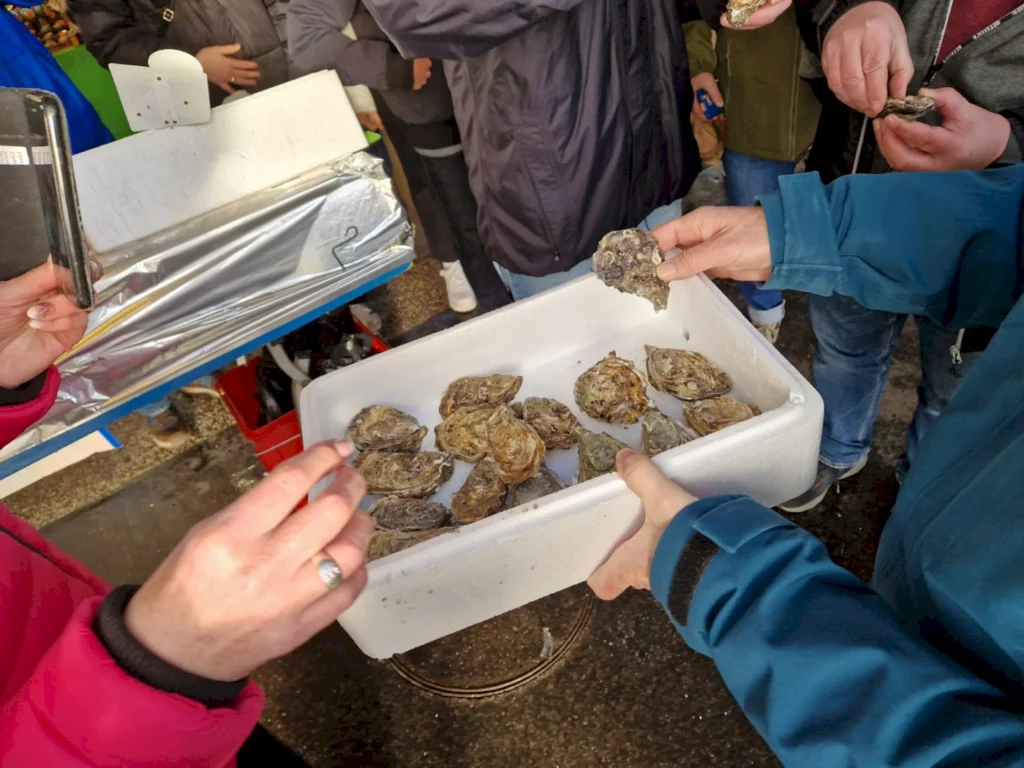 Menschen beim Kauf von frischen Austern an einem Marktstand, wobei eine Person eine Auster aus einem Behälter hält und eine andere Person ein Smartphone benutzt.