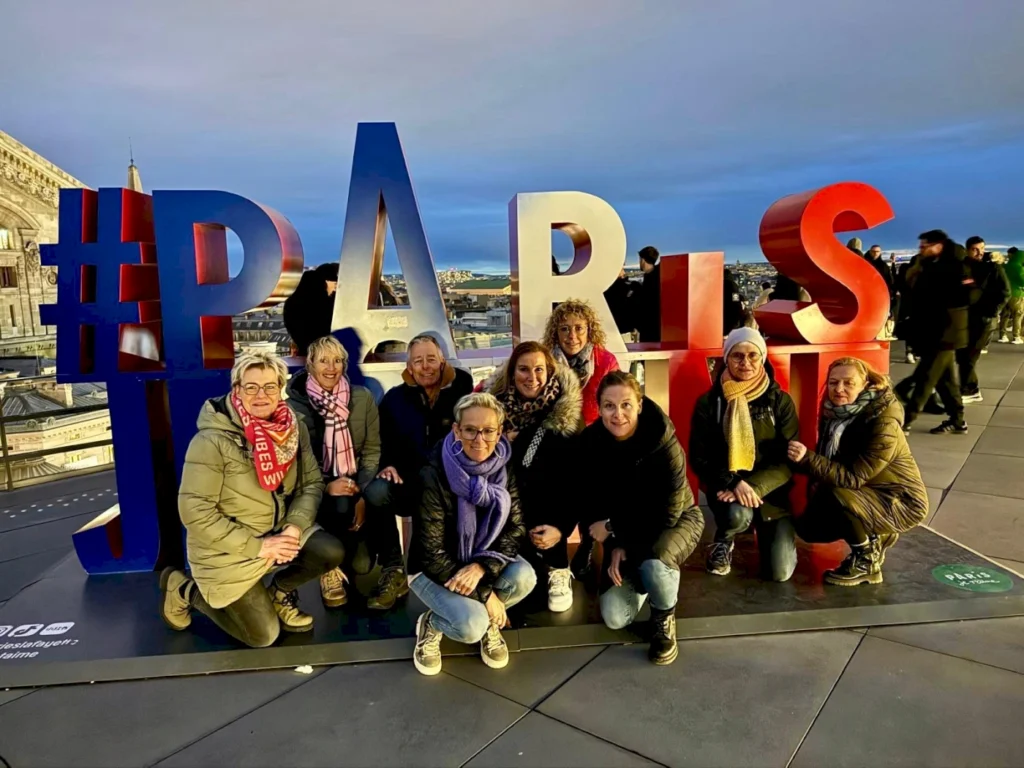 Gruppe von Frauen posiert vor dem bunten "PARIS"-Schild mit Hashtag auf einer Aussichtsplattform, umgeben von der Stadtlandschaft von Paris bei Sonnenuntergang.