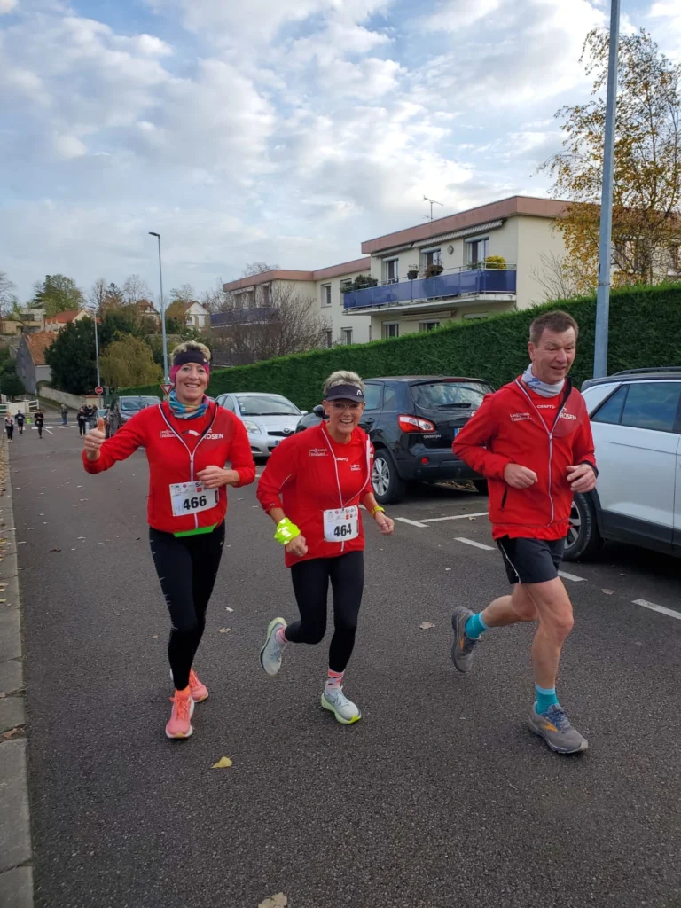 Drei Teilnehmer in roten Laufjacken nehmen an einem Straßenlauf teil, lächeln und zeigen Daumen hoch. Im Hintergrund sind Wohnhäuser und parkende Autos zu sehen.
