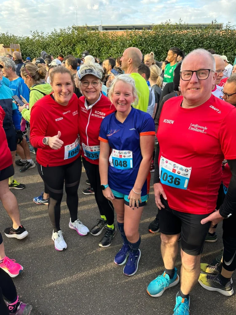 Vier Teilnehmer in Sportkleidung stehen lächelnd vor einer Menschenmenge bei einem Laufereignis. Sie tragen rote und blaue Shirts mit Startnummern und sind bereit für den Wettkampf.