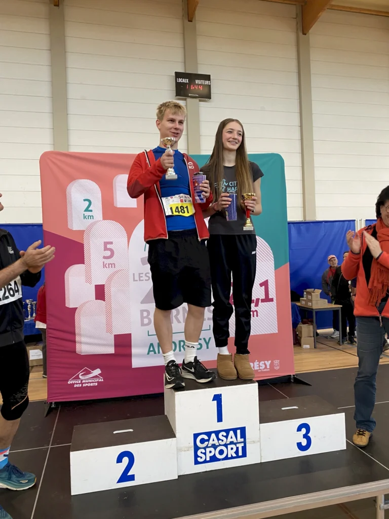Zwei junge Sportler stehen auf dem Siegertreppchen eines Laufwettbewerbs, halten Pokale in der Hand und feiern ihren Erfolg. Im Hintergrund sind Zuschauer zu sehen, während die Veranstaltung in einer Sporthalle stattfindet.