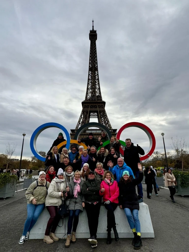 Gruppe von Menschen, die vor dem Eiffelturm in Paris posiert, umgeben von den Olympischen Ringen. Die Szene zeigt eine fröhliche Atmosphäre mit Teilnehmern in Winterkleidung und verschiedenen Gesten der Begeisterung.