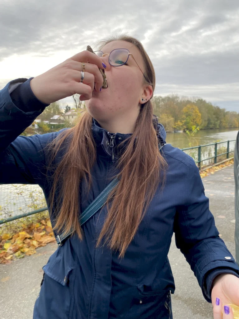 Junge Frau mit Brille, die am Ufer eines Gewässers eine Muschel isst, umgeben von herbstlicher Landschaft.