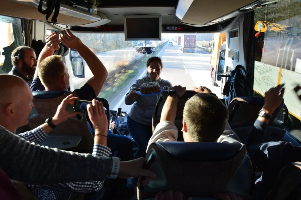 Gruppe von Passagieren in einem Bus, die eine Feier mit einem Kuchen und Kerzen genießen. Einige halten Smartphones, um den Moment festzuhalten, während die Fahrerin den Kuchen präsentiert. Sonnenlicht strömt durch die Fenster, während die Autobahn im Hintergrund sichtbar ist.
