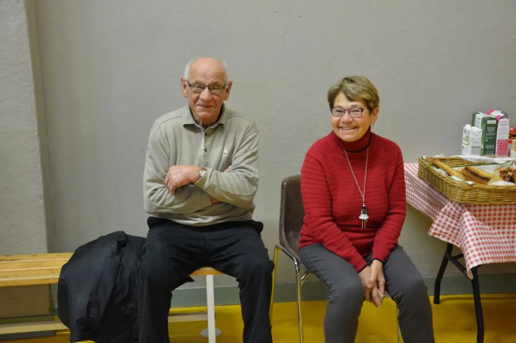 Ein älterer Mann und eine Frau sitzen nebeneinander auf einer Bank in einem Gemeinschaftsraum. Der Mann trägt ein graues Sweatshirt und hat eine Brille auf, während die Frau ein rotes Strickoberteil und eine Kette trägt. Im Hintergrund ist ein Tisch mit einem Korb und Getränkekartons zu sehen.