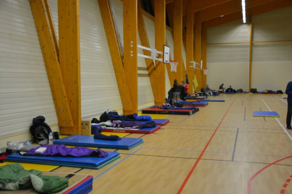 Innenansicht einer Turnhalle mit Schlafmatten und Schlafsäcken, die als Notunterkunft genutzt werden. Menschen sind mit persönlichen Gegenständen beschäftigt, während Basketballkörbe im Hintergrund sichtbar sind.