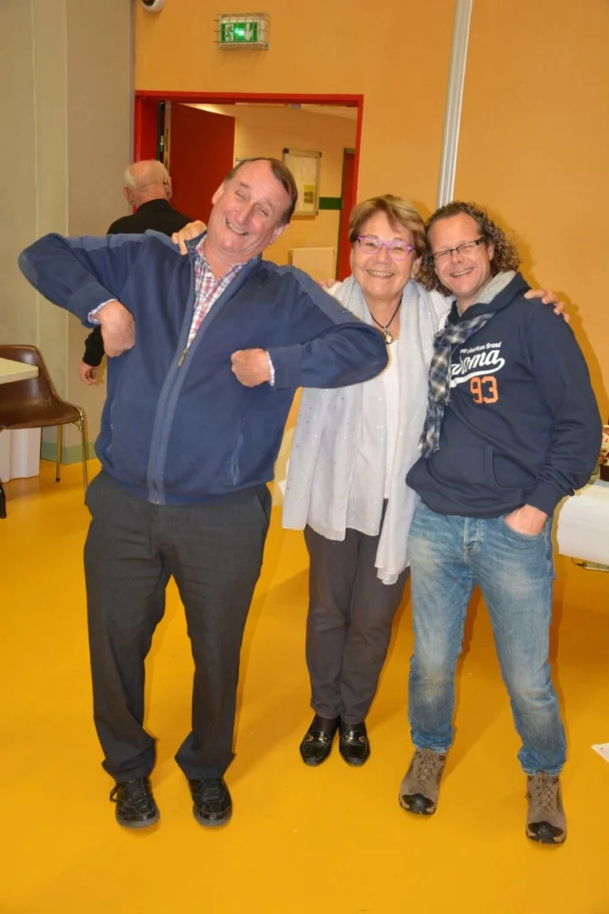 Drei Personen posieren fröhlich vor einem bunten Hintergrund. Ein Mann zeigt einen schelmischen Gesichtsausdruck, während die Frau in der Mitte lächelt und die andere Person rechts ebenfalls lächelt. Alle tragen legere Kleidung und scheinen eine angenehme Zeit zu haben.