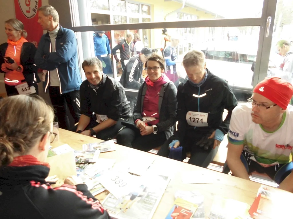 Teilnehmer eines Laufwettbewerbs sitzen an einem Tisch, während sie auf ihre Startnummern warten. Im Hintergrund sind weitere Sportler und ein Informationsstand zu sehen. Die Atmosphäre ist lebhaft und sportlich.