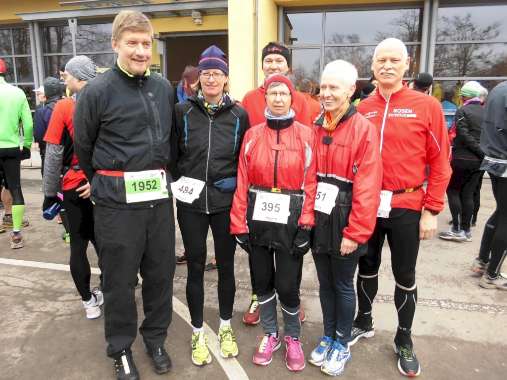 Gruppe von sechs Laufteilnehmern in Sportkleidung mit Startnummern, die sich vor einem Gebäude versammeln. Sie tragen wetterfeste Jacken und Laufhosen, bereit für ein Rennen bei kühlem Wetter.