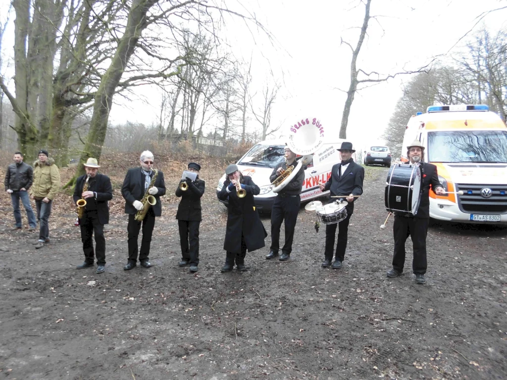 Eine siebenköpfige Blaskapelle spielt im Freien, umgeben von Bäumen und einem unbefestigten Weg. Im Hintergrund sind zwei Fahrzeuge, darunter ein Krankenwagen, sichtbar. Die Musiker tragen schwarze Anzüge und Hüte, während sie verschiedene Blasinstrumente und ein Schlagzeug spielen. Einige Zuschauer stehen in der Nähe und beobachten die Aufführung.