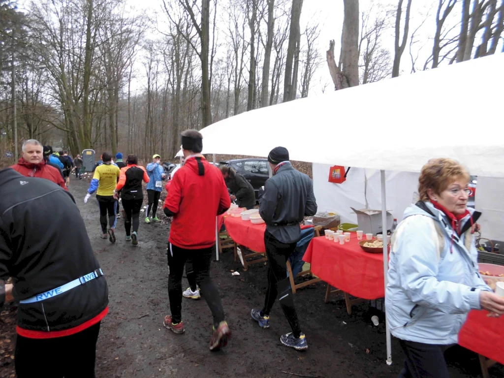 Läufer bei einem Wettkampf im Wald, vorbei an einem Verpflegungsstand mit Tischen und Snacks, während Zuschauer und Helfer im Hintergrund aktiv sind.