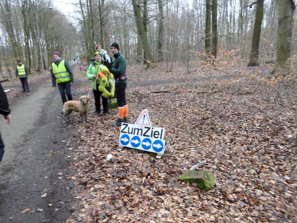 Gruppe von Menschen in Warnwesten steht auf einem Waldweg, während ein Hund daneben sitzt. Ein Schild mit der Aufschrift "Zum Ziel" und Richtungspfeilen zeigt den Weg. Herbstliche Umgebung mit Laub auf dem Boden.