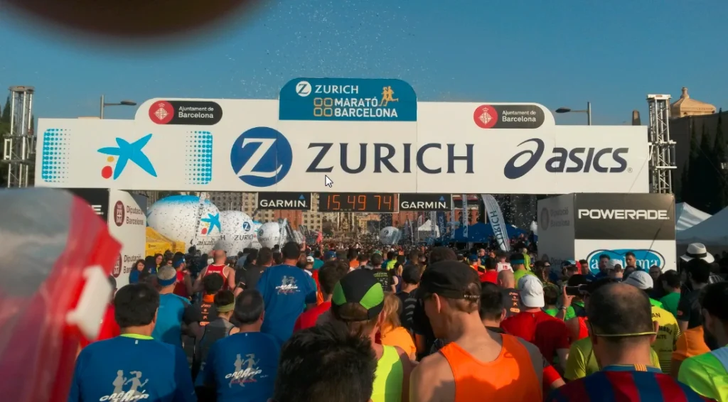 Startschuss für den Zurich Maratón de Barcelona mit Teilnehmern unter dem Zielbogen, umgeben von Sponsoren-Logos und jubelndem Publikum.