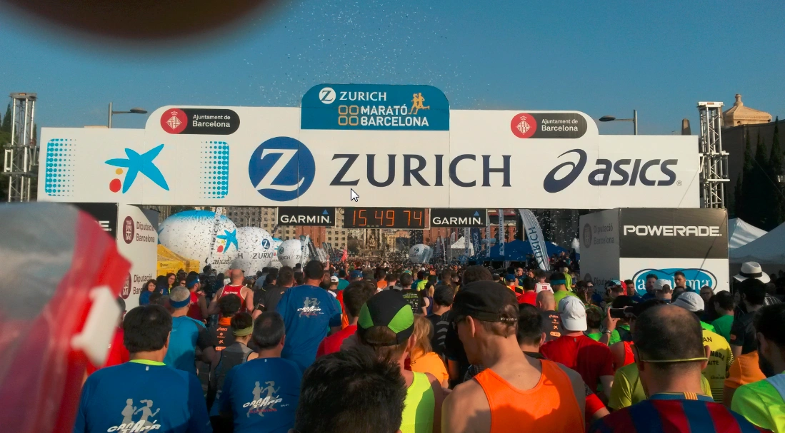 Startschuss für den Zurich Maratón de Barcelona mit Teilnehmern unter dem Zielbogen, umgeben von Sponsoren-Logos und jubelndem Publikum.