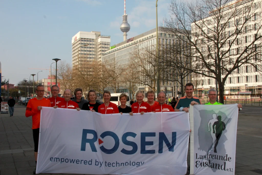 Gruppe von Läufern mit Rosen-Banner in einer Stadt, Fernsehturm im Hintergrund, sonniger Tag.