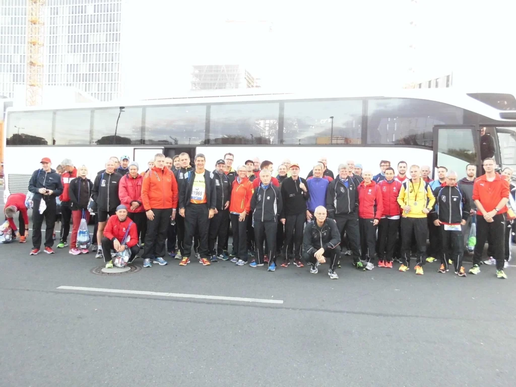 Gruppe von Läufern vor einem Reisebus, bereit für ein Event in der Stadt, alle in Sportkleidung und teils mit Startnummern.