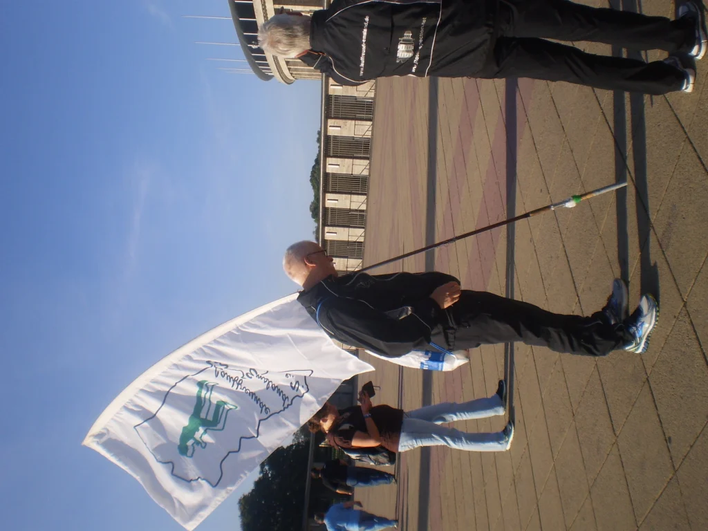 Person trägt auf dem Platz vor einem großen Gebäude eine Flagge mit grüner Figur, bei sonnigem Wetter.