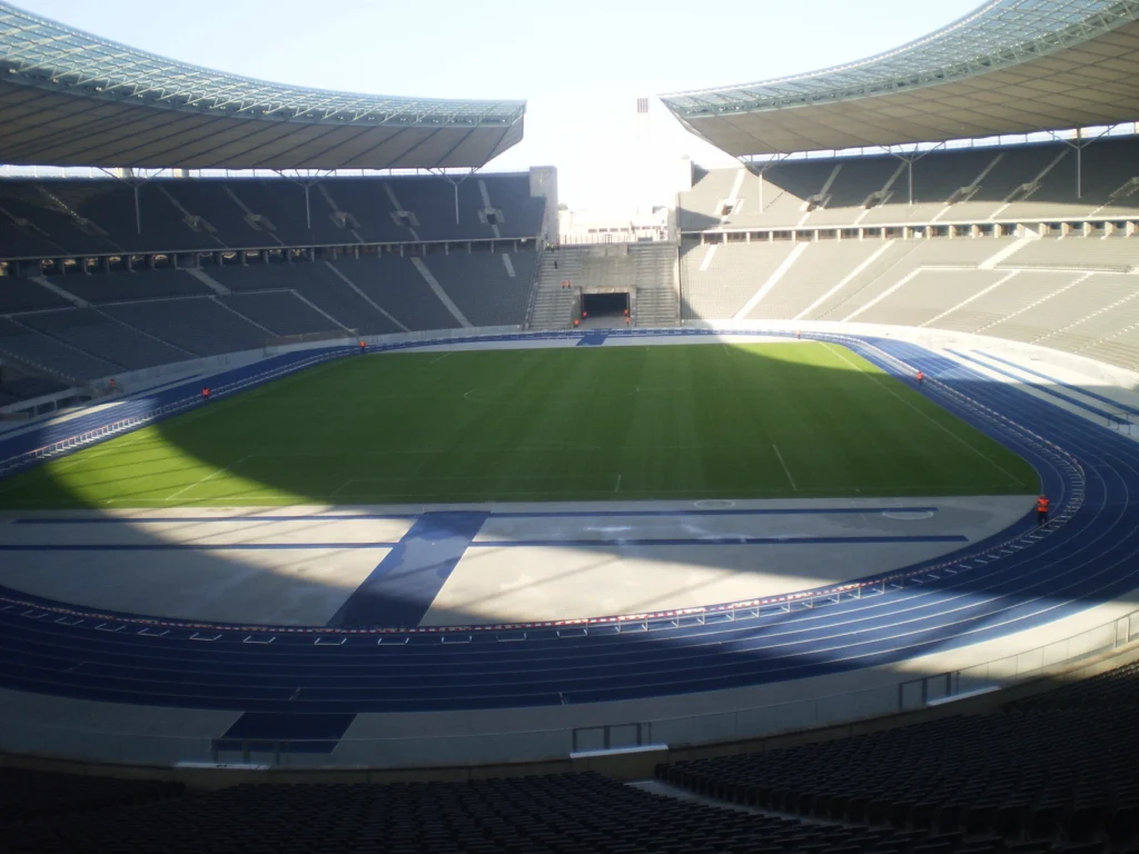Leeres Stadion mit grünem Spielfeld und blauer Laufbahn, modern und architektonisch beeindruckend.