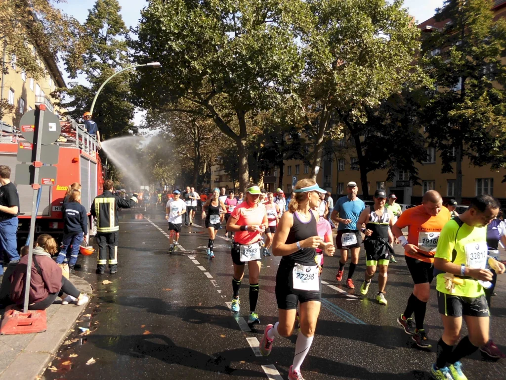 Läufer beim Stadtmarathon, Feuerwehr sprüht Wasser zur Abkühlung, sonniger Tag, grüne Bäume, Zuschauer am Straßenrand.