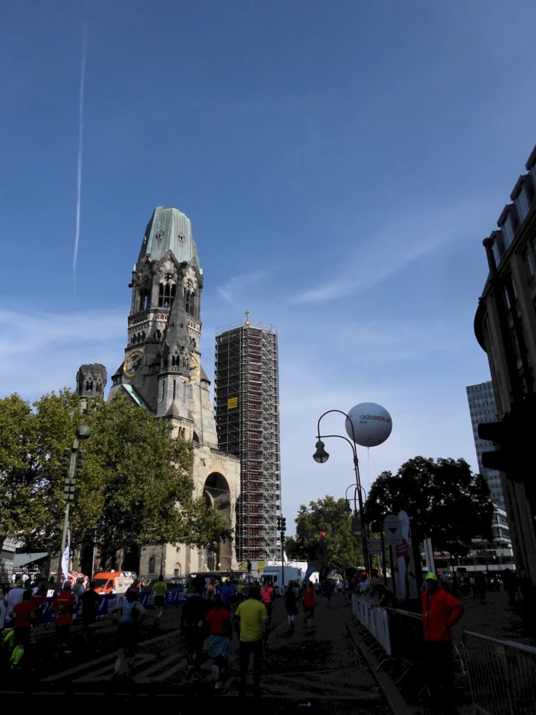 Kaiser-Wilhelm-Gedächtniskirche in Berlin mit Baugerüst und vorbeilaufenden Menschen bei sonnigem Wetter.