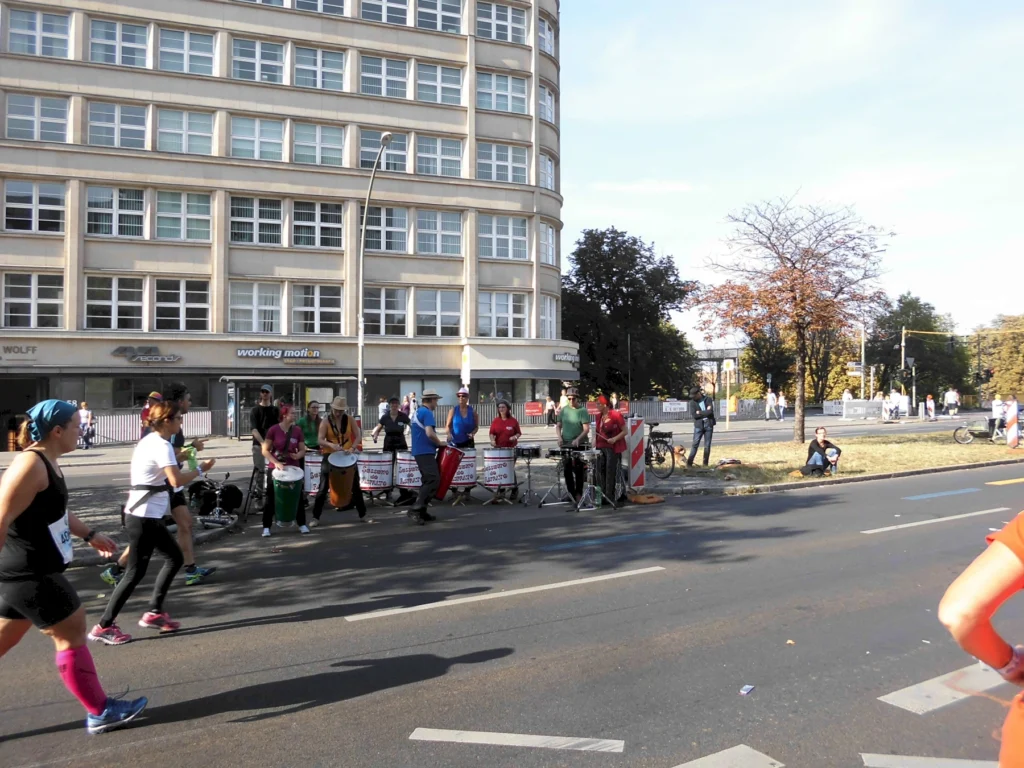 Läufer beim Straßenlauf, begleitet von Trommlern vor städtischer Kulisse, unterstützen das Event mit Rhythmen.